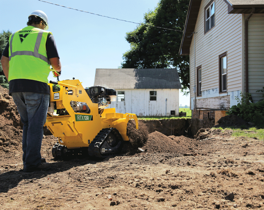 A man with a reflective jacket on pushes a walk-behind trencher as it trenches in a residential yard