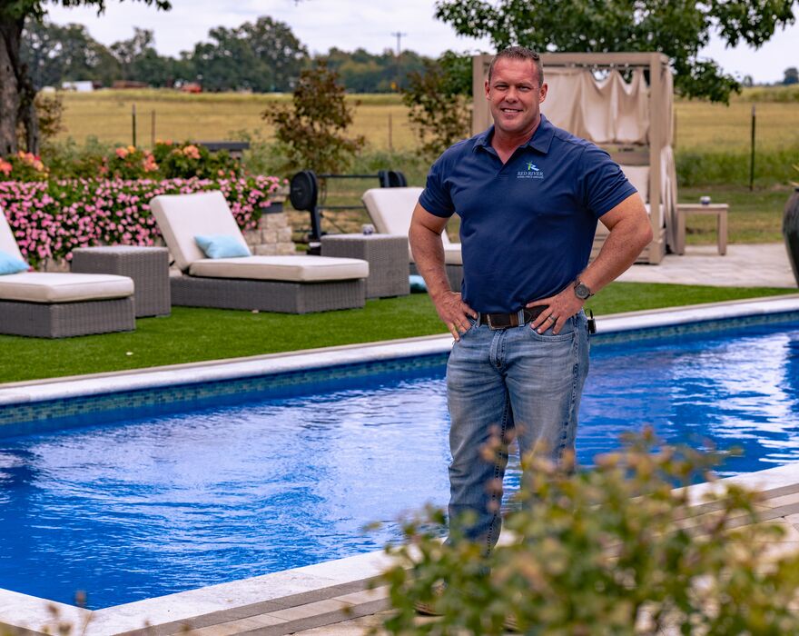 A man in a polo and jeans stands in front of a well manicured pool