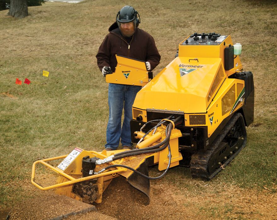 A man operates a stump grinder machine from a panel next to the machine