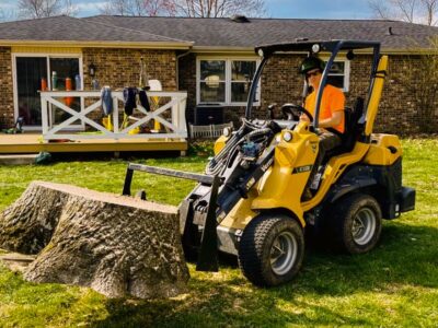 A man sits in a compact articulated loader and lifts a stump