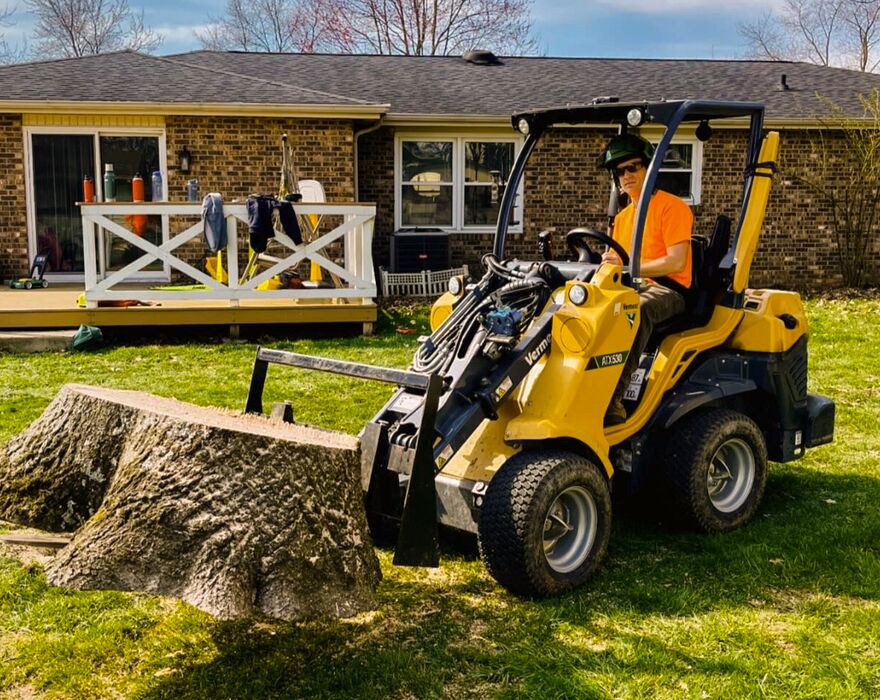 A man sits in a compact articulated loader and lifts a stump