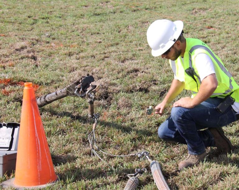 A man wearing a hard hat and a yellow reflective jacket crouches on the ground near an orange cone.