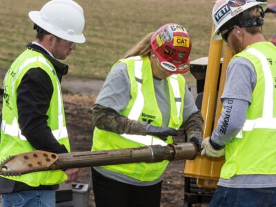 Three people in hardhats and reflective vests inspect a piece of horizontal directional drill tooling.