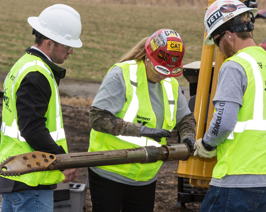 Three people in hardhats and reflective vests inspect a piece of horizontal directional drill tooling.