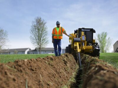 : A man in an orange reflective vest stands next a small yellow trencher as it digs into the ground.