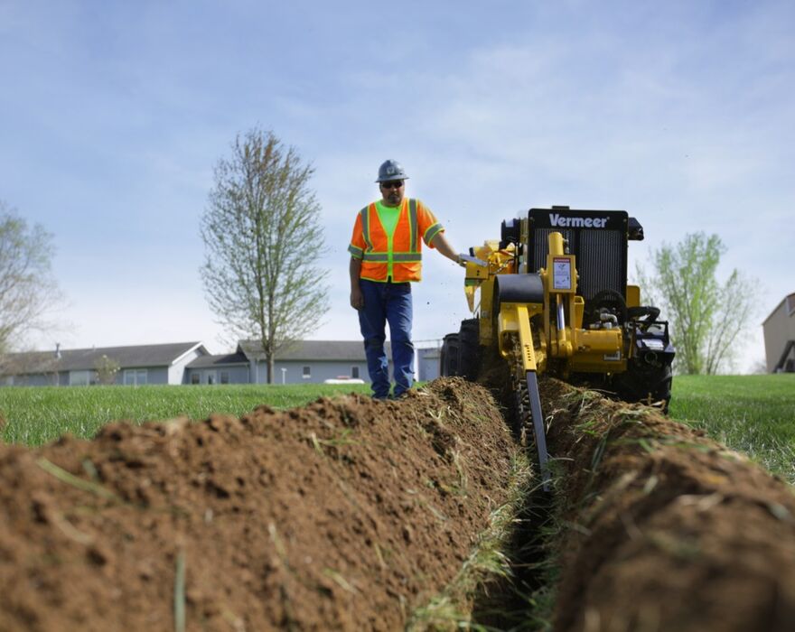 : A man in an orange reflective vest stands next a small yellow trencher as it digs into the ground.