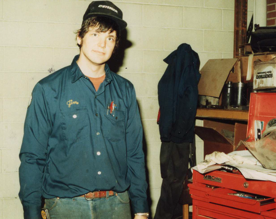 A photo from the 1980s of a man in a shop with a hat on and a shirt that says "Jim"