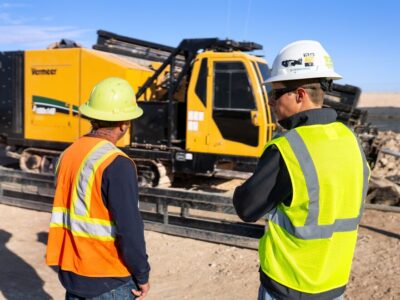 Two men stand in front of a pipeline horizontal directional drill