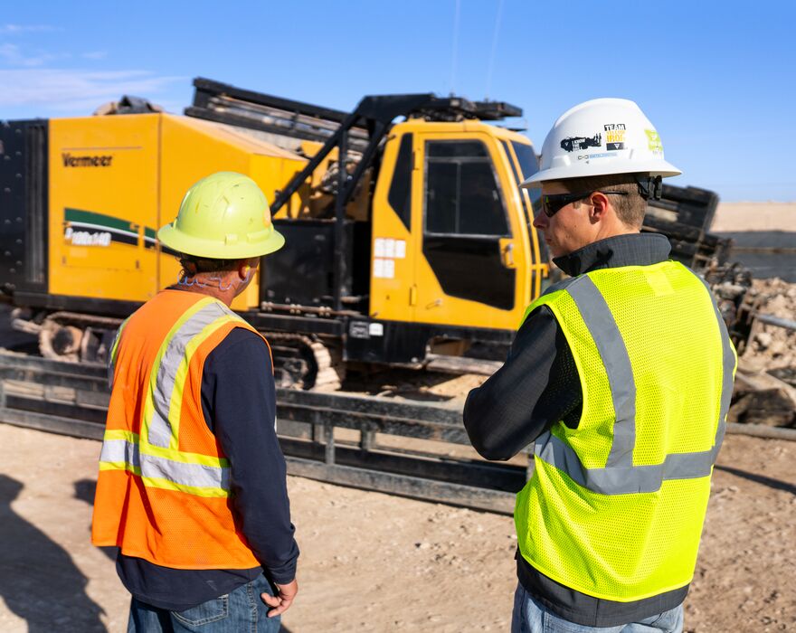 Two men stand in front of a pipeline horizontal directional drill